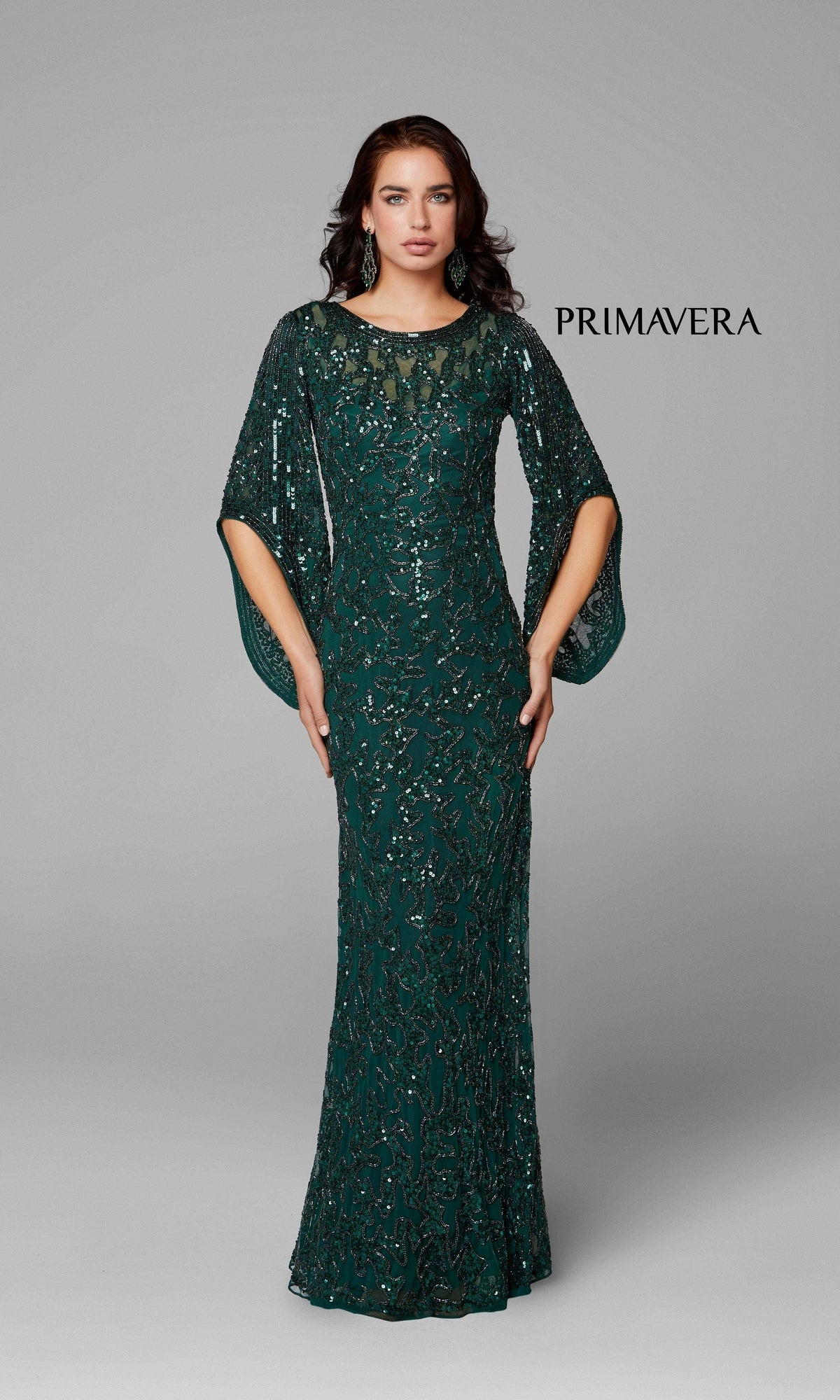 A woman poses against a plain gray background in the Long Bell Sleeve Beaded Evening Gown 9713—an emerald green, floor-length dress with beading and sheer sleeves. The word